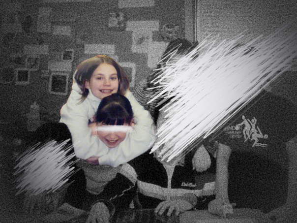 black and white vignette image of 5th grade Shannon smiling joyfully and hugging Rachel, whose face is scratched out to conceal her identity, in a classroom. They are surrounded by other classmates, whose faces are also scratched out to protect their identities.