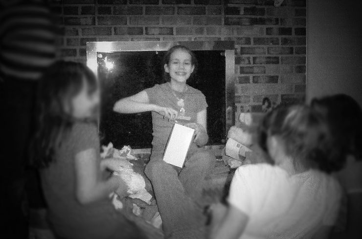 blurry, grainy black and white photo of young Shannon sitting in front of a fireplace opening a present surrounded by four of her friends who are facing her